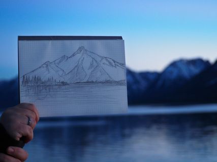 a hand holding up a sketch to match mountains behind it